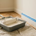 Rouleau chargé de peinture blanc cassé dans un bac gris, avec ruban de masquage bleu sur la plinthe d’une chambre